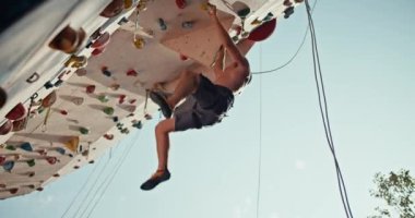 Bottom view: an athletic guy in black shorts with a sculpted torso climbs up the climbing wall and hangs on the ledge of the training route of the climbing wall on a sunny summer day.