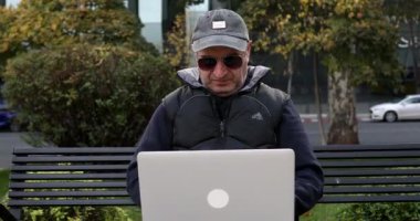 Kendine güvenen orta yaşlı bir adam bankta oturuyor ve sokakta dizüstü bilgisayar kullanıyor.