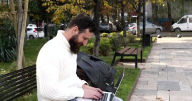 Dışarıda dizüstü bilgisayarla çalışan sakallı genç bir adam. Şehir caddesinde dizüstü bilgisayarda yazan yakışıklı adam..