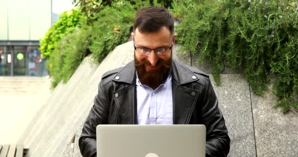 ラップトップコンピュータの屋外で働いている髭の若い男 街の通りにノートパソコンのキーボードを入力するハンサムな男 — ストック動画