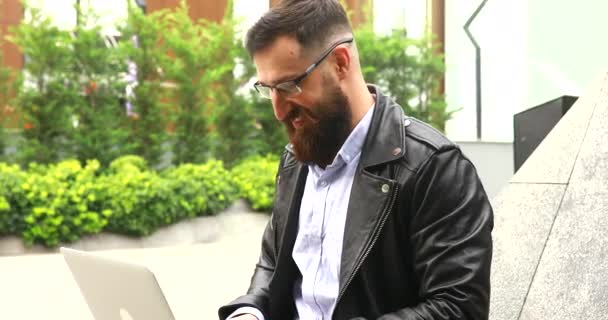 ラップトップコンピュータの屋外で働いている髭の若い男 街の通りにノートパソコンのキーボードを入力するハンサムな男 — ストック動画