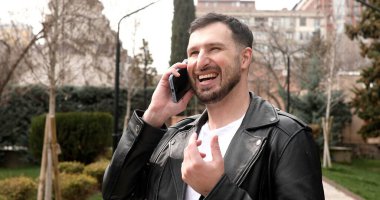 Gülümseyen genç adam dışarıda cep telefonuyla konuşuyor.