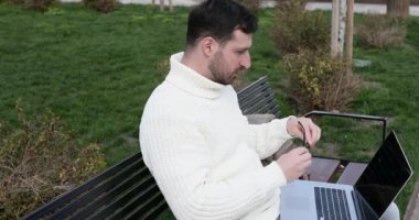 Yakışıklı beyaz adam bankta oturuyor, dizüstü bilgisayar kullanıyor ve göz ağrısı çekiyor.