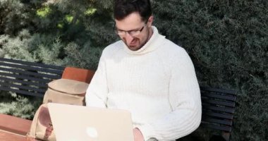 Genç adam bankta oturuyor ve parkta dizüstü bilgisayar kullanıyor.