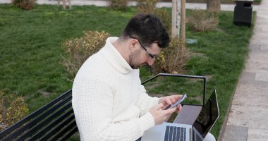 Genç beyaz adam bankta oturuyor, dışarıda dizüstü bilgisayar ve akıllı telefon kullanıyor.