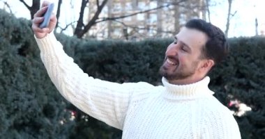 Yakışıklı, mutlu, genç bir adam akıllı telefon tutuyor ve sokakta selfie çekiyor.