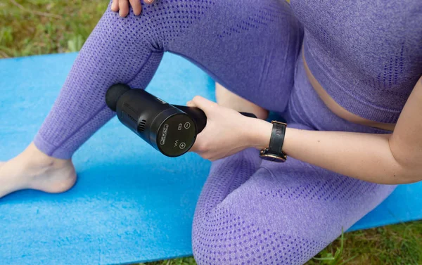 年轻健康的女人坐在瑜伽垫上 用撞击按摩器按摩室外肌肉 — 图库照片