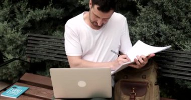 Odaklanmış genç adam bankta oturuyor, dizüstü bilgisayar kullanıyor ve parkta defter yazıyor.
