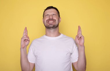Beyaz tişörtlü genç adam sarı arka planda parmaklarını çapraz gösteriyor..