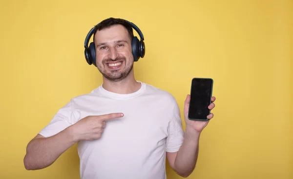 happy smiling man in headphones showing smartphone isolated over yellow background