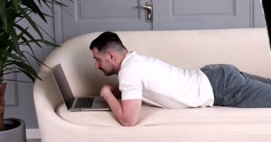 Sakallı yakışıklı genç adam kanepede uzanıyor ve evde dizüstü bilgisayar kullanıyor..