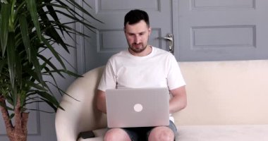 Sakallı yakışıklı genç adam kanepede oturuyor ve evde dizüstü bilgisayar kullanıyor..