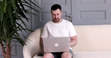 Sakallı yakışıklı genç adam kanepede oturuyor ve evde dizüstü bilgisayar kullanıyor..