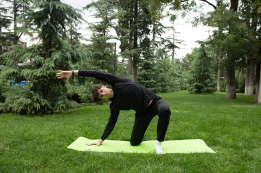 Güneşli bir sabahta parkta yoga yapan bir adam. Açık havada yoga minderinde egzersiz yapan sağlıklı ve aktif yaşam tarzı kavramı olan zinde bir genç. Açık havada eğitim..