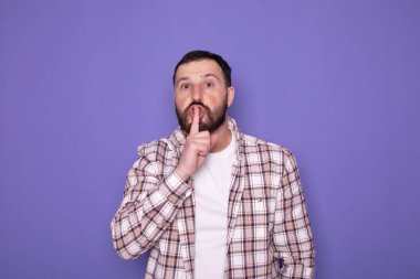 Yakışıklı sakallı adam mor arka planda sessizlik jesti yapıyor.