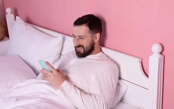 Bearded young man in pajama using mobile phone on bed.