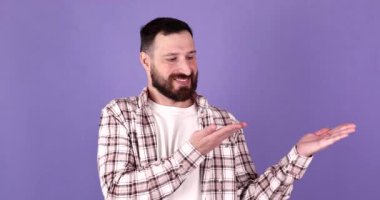 Genç beyaz sakallı yakışıklı adam elleriyle bir şey sunuyor, bir ürün gösteriyor, mor arka planda gülümsüyor ve neşeli..