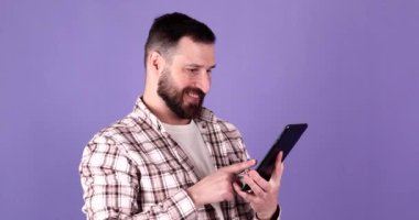 Genç sakallı adam mor arka planda dijital tablet kullanıyor..