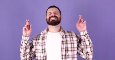 Genç sakallı adam dilek diliyor parmaklarını mor arka plan stüdyo portresinde izole halde tut.. 