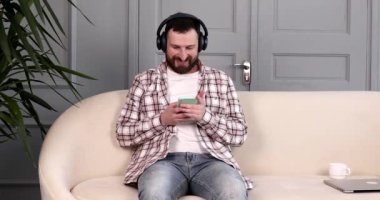 Neşeli genç sakallı adam kanepede dinleniyor ve akıllı telefon uygulaması kullanıyor, evde kanepede dinlenirken kulaklıkla müzik dinliyor..