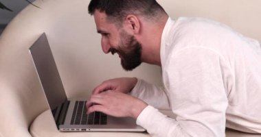 Yatak odasında dizüstü bilgisayar kullanan pijamalı genç sakallı bir adam..