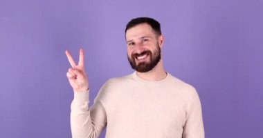 Beyaz sakallı adam Ziyaretçi işareti gösterirken neşeli ve mutlu görünüyor. Mor arka planda zafer için jest yapan gülümseyen bir adamın portresi..