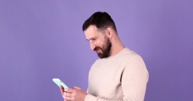 Mutlu, sakallı, sıradan kıyafetleri olan, cep telefonu kullanan mutlu adam evet diyor, mor stüdyo arka planında kazanan hareketi yapıyor..