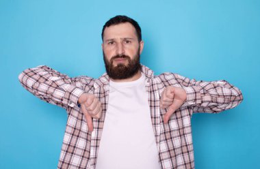 Mavi arka planda baş parmakları aşağıda olan tatmin olmamış sakallı bir adamın portresi. Stüdyo çekimi.