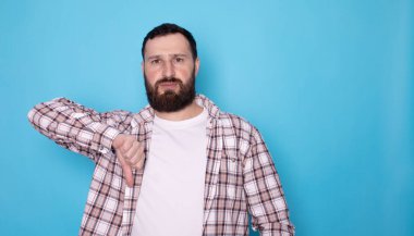 Mavi arka planda baş parmakları aşağıda olan tatmin olmamış sakallı bir adamın portresi. Stüdyo çekimi.