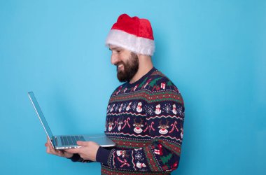 Sakallı bilişim teknolojisi uzmanı örgü kazak ve Noel Baba şapkası takıyor. Mavi arka planda dizüstü bilgisayar kullanıyor..     
