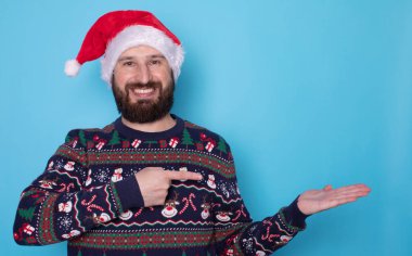 Noel süveteri ve açık mavi arka planda bir şeye bakan Noel Baba şapkalı mutlu genç adam. Metin için boşluk.    