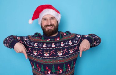 Noel süveteri ve açık mavi arka planda bir şeye bakan Noel Baba şapkalı mutlu genç adam. Metin için boşluk.    