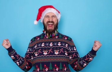 Yeni yılı kutlayan, süveter ve Noel Baba şapkası giyen, sevinç çığlıkları atan, kazanan ya da zafer kazanan, mavi arka planda dikilen duygusal ve çekici sakallı bir adamın portresi..