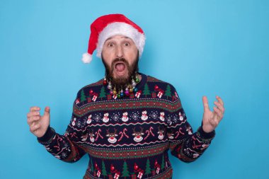 Yeni yılı kutlayan, süveter ve Noel Baba şapkası giyen, sevinç çığlıkları atan, kazanan ya da zafer kazanan, mavi arka planda dikilen duygusal ve çekici sakallı bir adamın portresi..