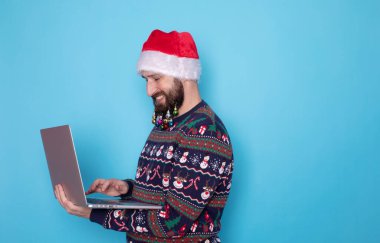 Sakallı bilişim teknolojisi uzmanı örgü kazak ve Noel Baba şapkası takıyor. Mavi arka planda dizüstü bilgisayar kullanıyor..     