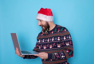 Sakallı bilişim teknolojisi uzmanı örgü kazak ve Noel Baba şapkası takıyor. Mavi arka planda dizüstü bilgisayar kullanıyor..     