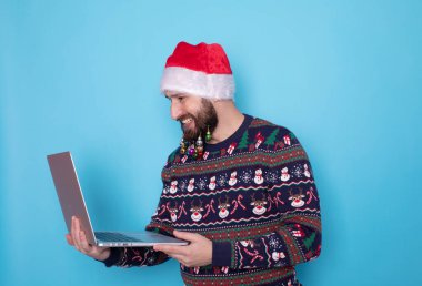Sakallı bilişim teknolojisi uzmanı örgü kazak ve Noel Baba şapkası takıyor. Mavi arka planda dizüstü bilgisayar kullanıyor..     