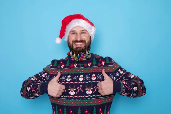 クリスマスのセーターとサンタの帽子に明るい青の背景に親指を示す幸せな若いひげ付き男 — ストック写真