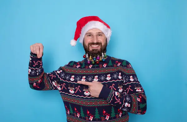 クリスマスセーターでハッピーな若いひげ付き男とサンタの帽子は ライトブルーの背景に何かを指している テキスト用スペース — ストック写真