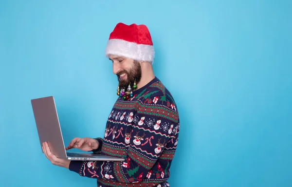 身穿针织毛衣 头戴圣诞帽 留着胡子的It人员 手持笔记本电脑 用蓝色背景隔离 — 图库照片