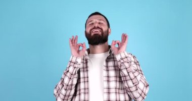 Soyutlanmış mavi arka planda duran genç yakışıklı adam rahatla ve gülümse gözleri kapalı parmaklarıyla meditasyon hareketi yapıyor..