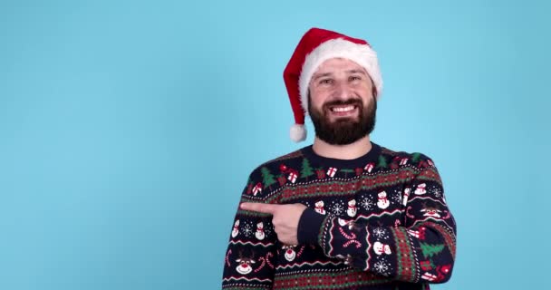 穿着圣诞毛衣头戴圣诞礼帽的快乐的小胡子男人指着浅蓝色背景的东西 案文的篇幅 — 图库视频影像