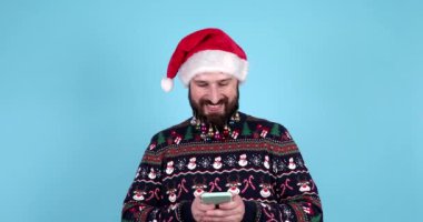 Sakallı adam Noel Baba şapkası takıyor. El ele tutuşup duruyor. Mavi arka planda izole edilmiş cep telefonu daktilo mesajı kullanıyor. Mutlu yıllar 2024 Noel tatili konsepti.