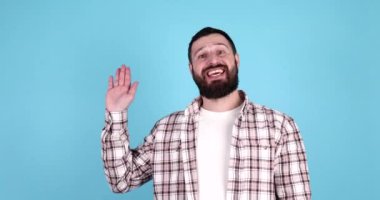 Genç, sakallı, sıradan bir gömlek giyen, mutlu ve gülümseyen, mavi arka planda dostça bir hoş geldin jesti yapan genç adam..