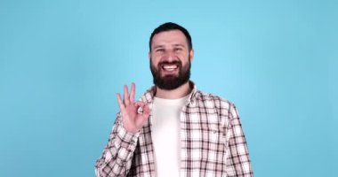 Mavi arka planda yalnız poz veren, güler yüzlü, sakallı, genç, yakışıklı bir adam. Tamam işareti gösteriliyor.