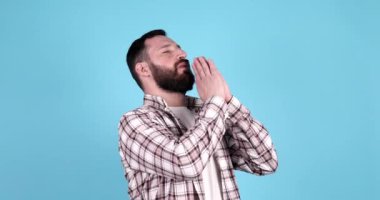 Mavi arka planda izole edilmiş genç sakallı beyaz adam el ele tutuşup dua ederken kendine güveniyor..