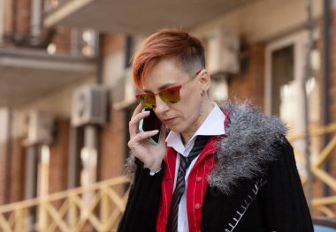 Kırmızı takım elbiseli, güneş gözlüklü, orta yaşlı güzel bir kadın akıllı telefondan sokağa karşı konuşuyor..