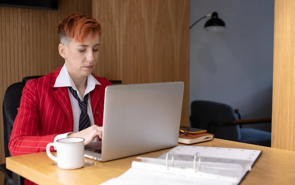 穿着红色西服的漂亮的成熟女商人在她的工作站上用笔记本电脑工作的肖像 — 图库照片