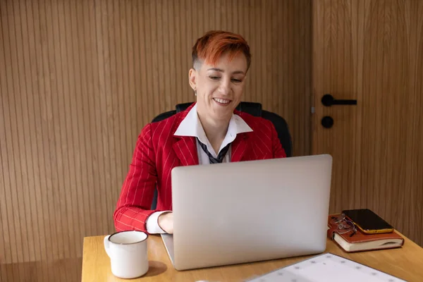 穿着红色西服的漂亮的成熟女商人在她的工作站上用笔记本电脑工作的肖像 — 图库照片