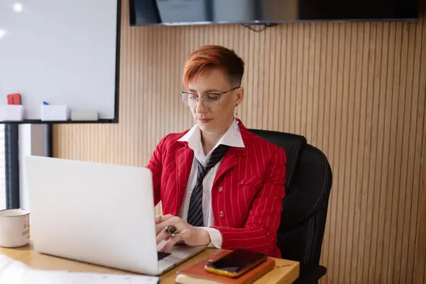 穿着红色西服的漂亮的成熟女商人的肖像和在她工作站的笔记本电脑上工作的眼镜 — 图库照片